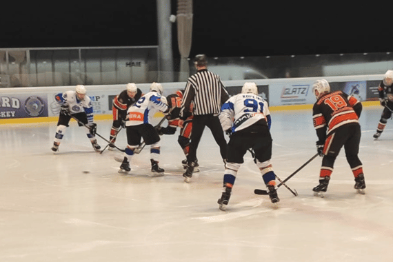 Eishockeyspiel mit sechs Spielern in Aktion; Schiedsrichter bereit für Puck-Einwurf; Hintergrund mit Werbebannern.