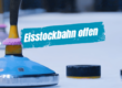 Eisstock auf Eisfläche mit Beschriftung "Eisstockbahn offen" bei eisiger Kulisse in Lindenberg.
