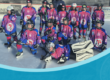Eine Gruppe Kinder in Eishockey-Ausrüstung posiert auf einer Eisfläche unter einem blauen Banner.