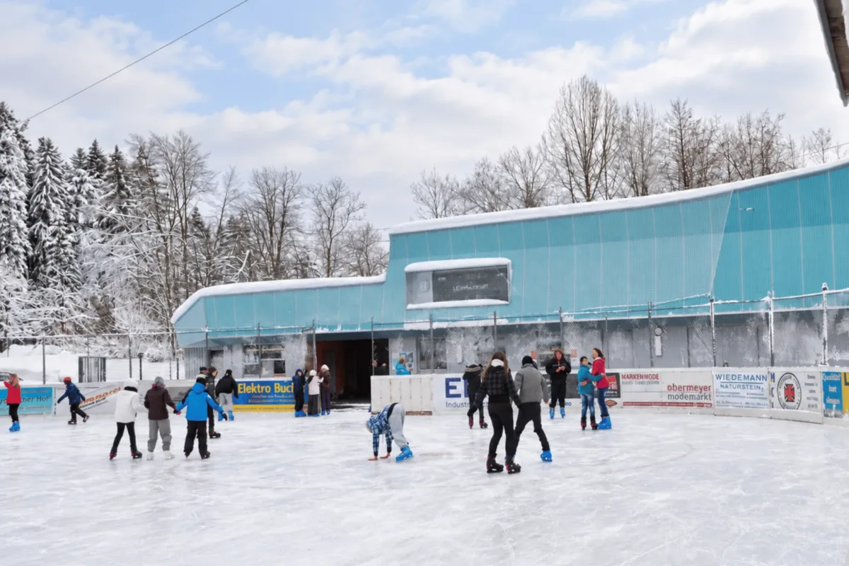 Eisläufer auf der schneeumrahmten Freiluft-Eisfläche des Eisplatz Lindenberg vor dem türkisen Stadiongebäude.