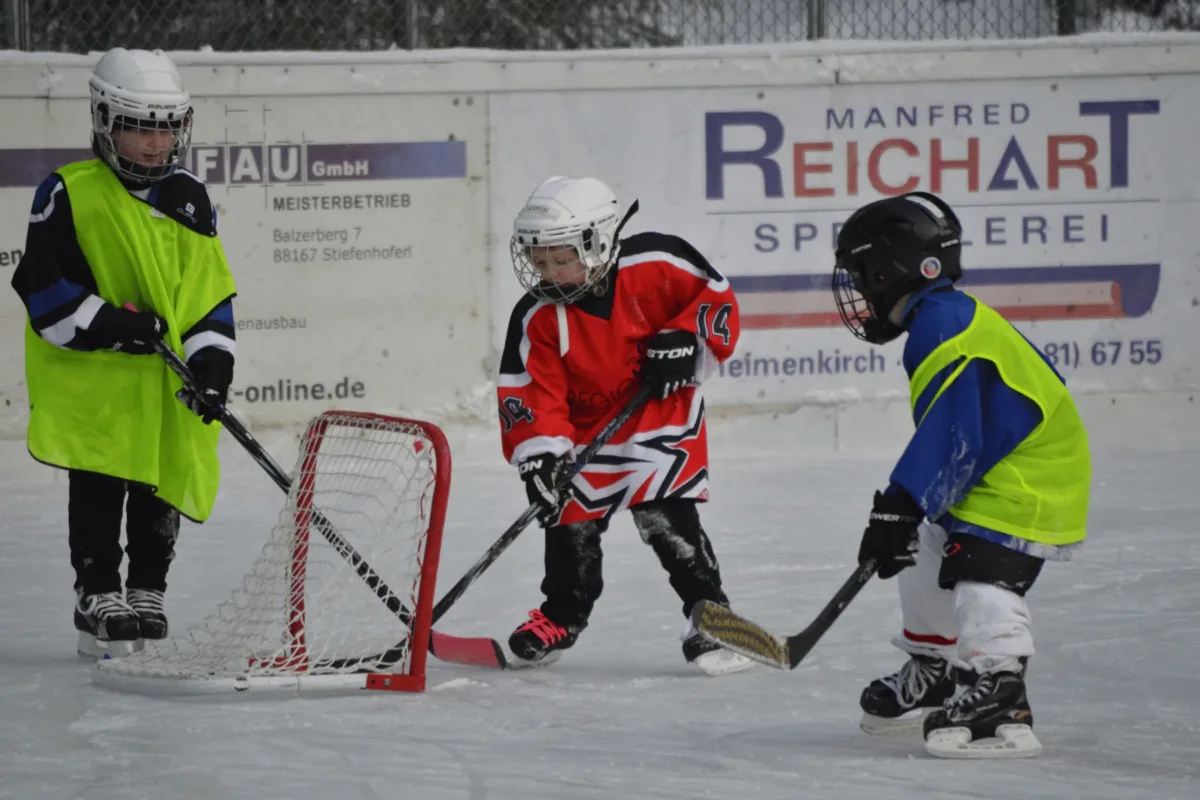 Drei Kinder in Eishockeyausrüstung spielen beim Nachwuchstraining am Eisplatz Lindenberg vor einem kleinen Tor.