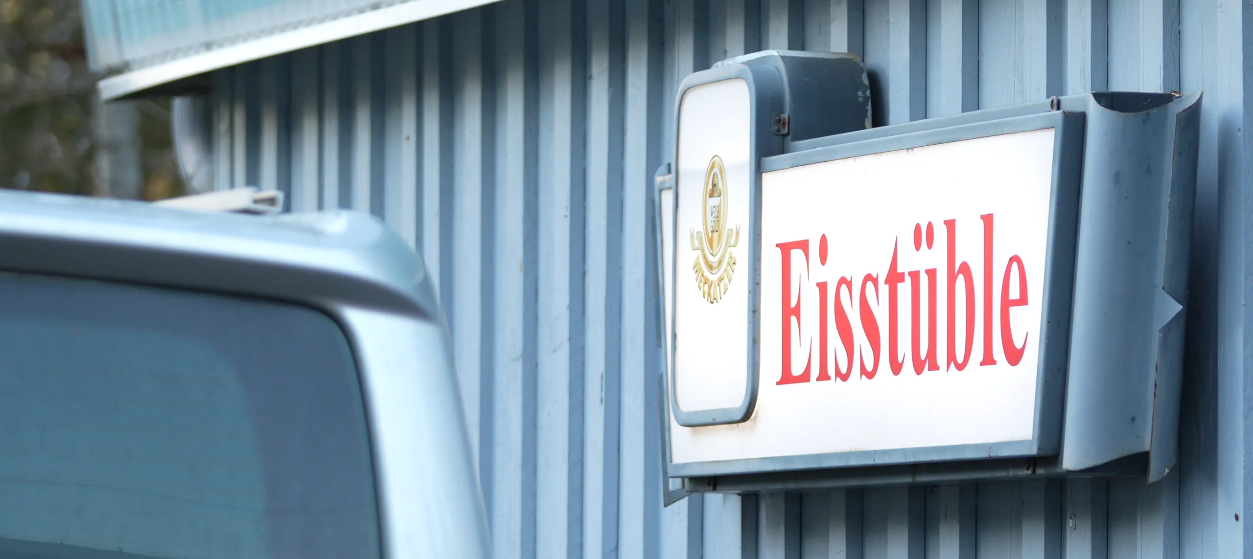 Leuchtschild „Eisstüble“ an der blauen Holzfassade des Eisplatz-Hauptgebäudes in Lindenberg