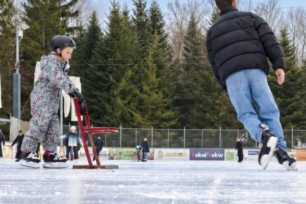 Ein Kind mit Lernhilfe und ein Erwachsener beim Eislaufen auf dem Eisplatz Lindenberg, im Hintergrund Tannen und Banden.