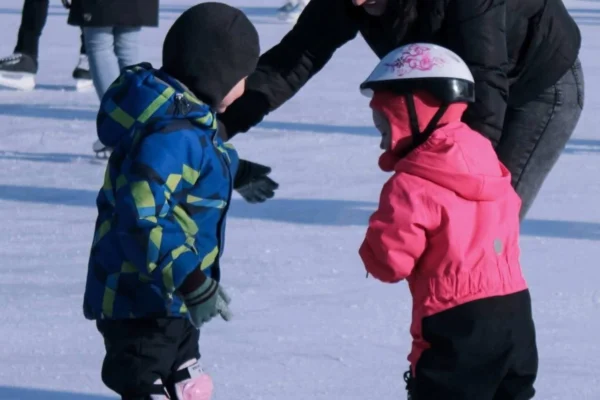 Zwei kleine Kinder mit Helm machen in der Eislaufschule Lindenberg ihre ersten Schritte auf dem Eis, eine Trainerin hilft.