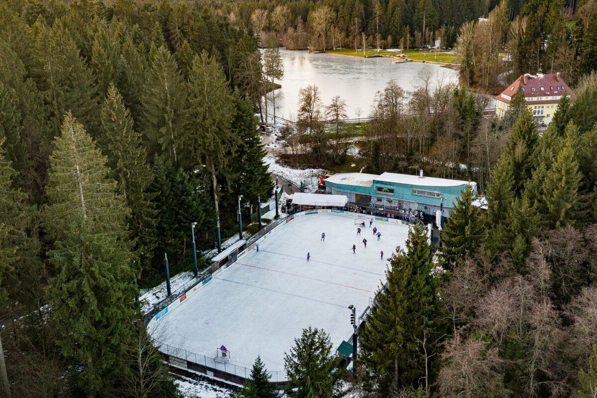 Luftaufnahme des Eisplatz Lindenberg: Eisfläche mit Läufern im Tannenwald, dahinter das Tribünengebäude und der Waldsee